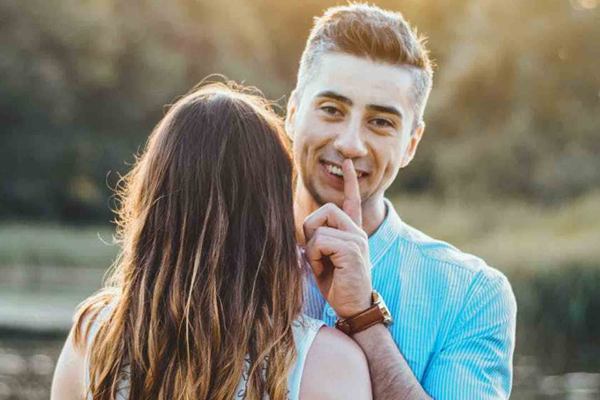 A man in a blue shirt holding an engagement ring behind her girlfriend.